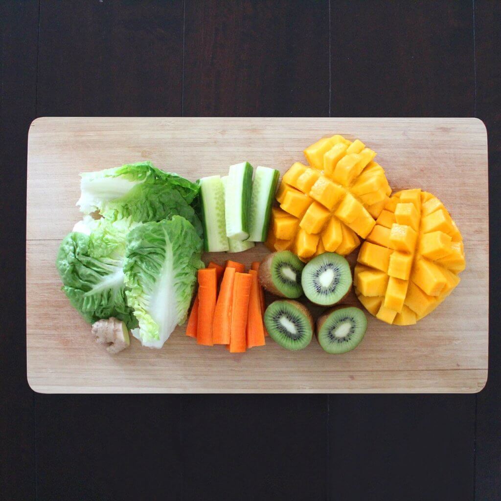 View from above of cutting board with chopped lettuce, cucumber, carrot, kiwi, and mango
